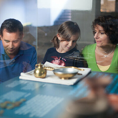 Familie hockt vor einer Beschreibungstafel für Badeutensilien in der Römerzeit