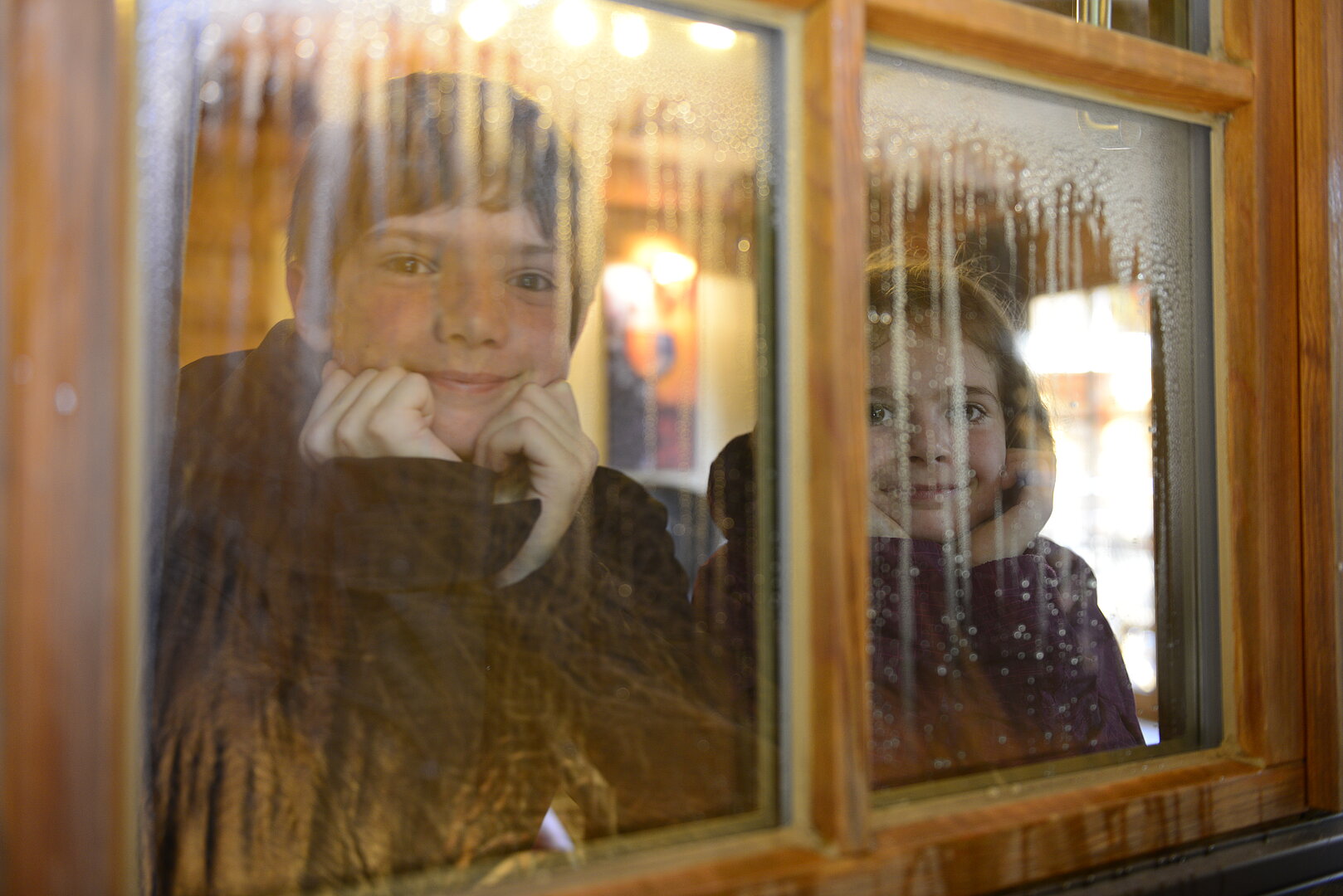 Mädchen und Junge gucken von drinnen durch Fensterscheibe nach draußen