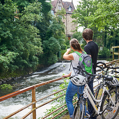 Ein Paar steht mit den Rädern an einem Flusslauf und schaut auf eine Burg